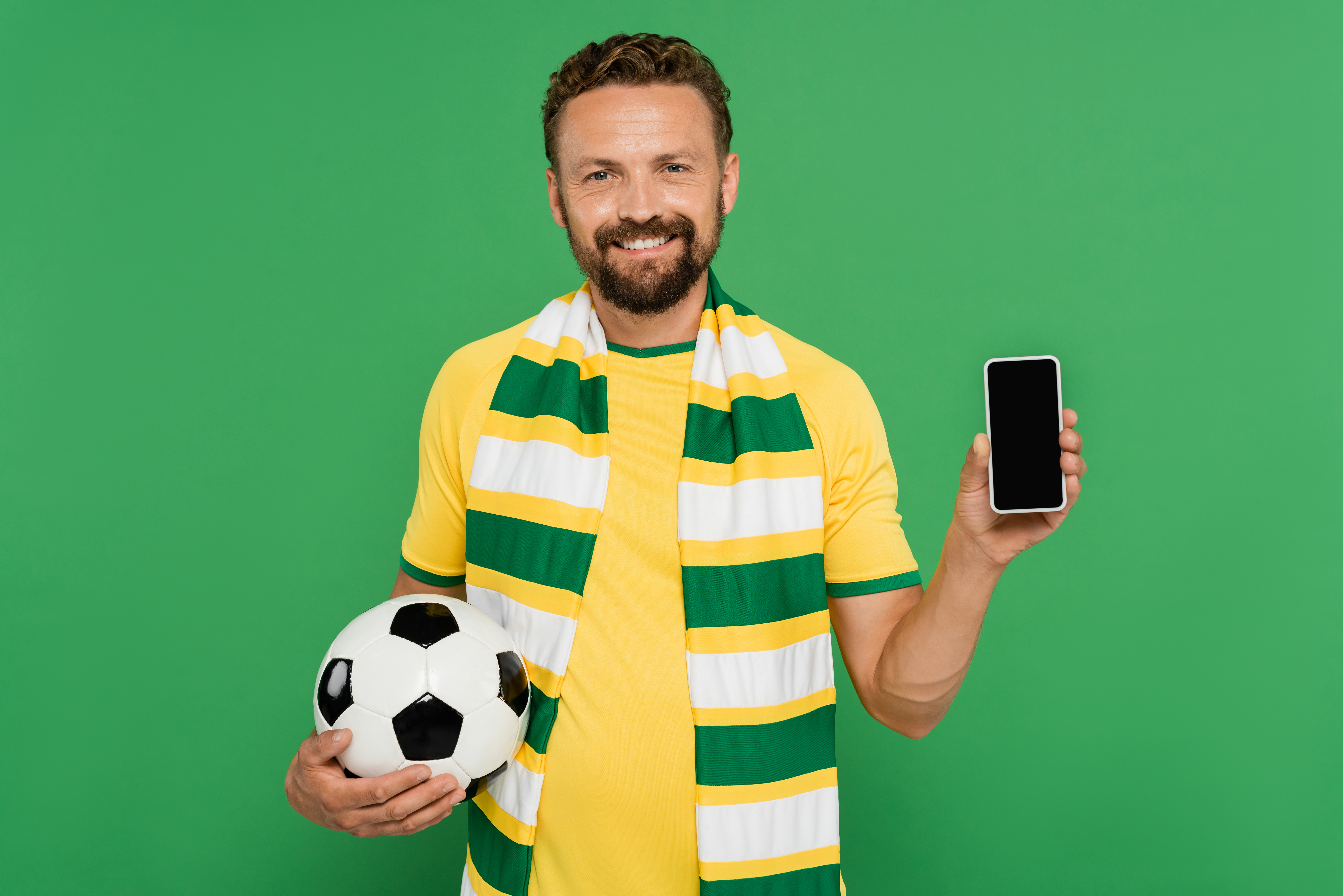 Homem alegre em cachecol listrado de verde amarelo e branco segurando bola de futebol e celular usado para apostar com pix no Brasil