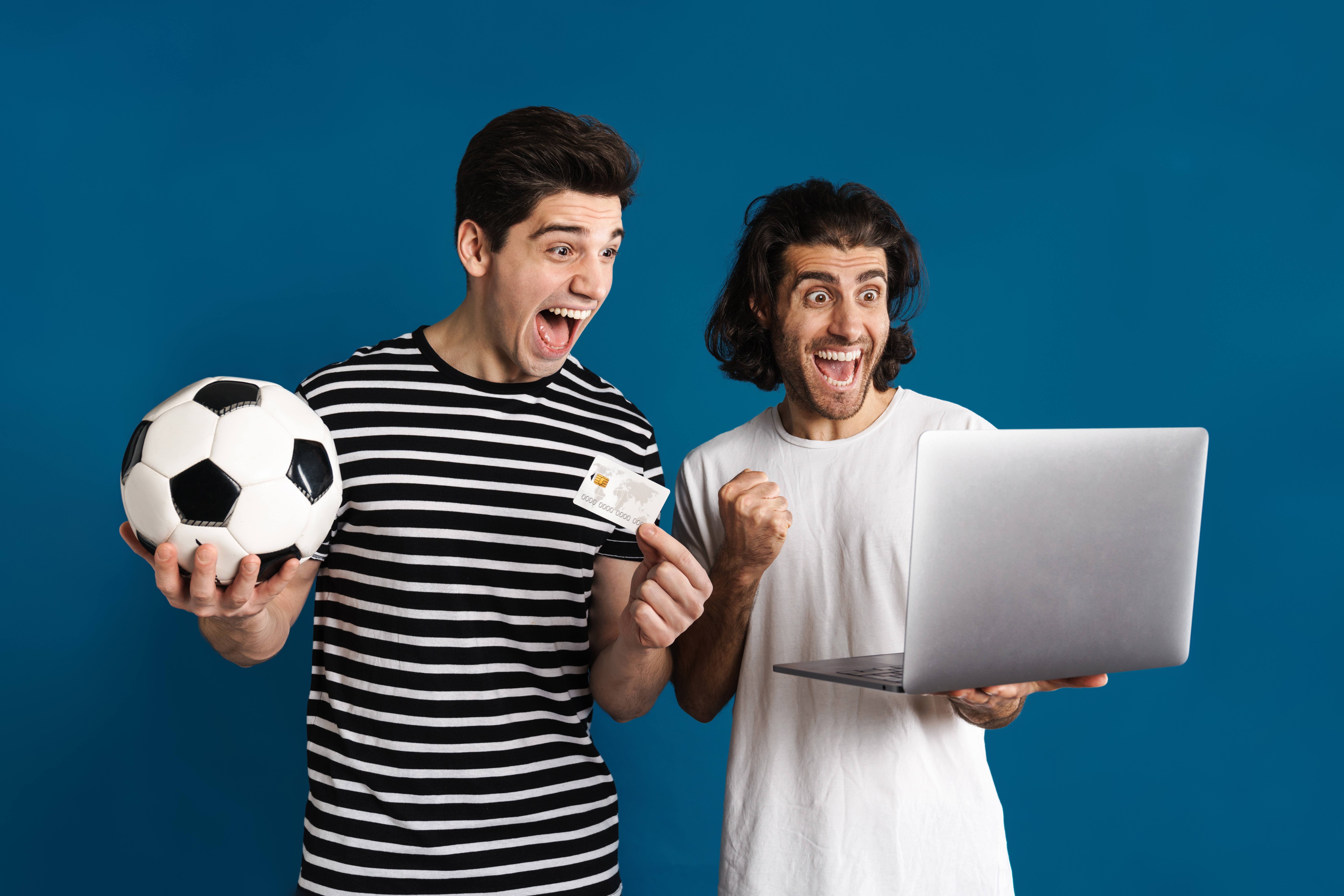 Dois homens animados usando laptop enquanto seguram bola de futebol e cartão de crédito para jogar em casas de apostas com cash out