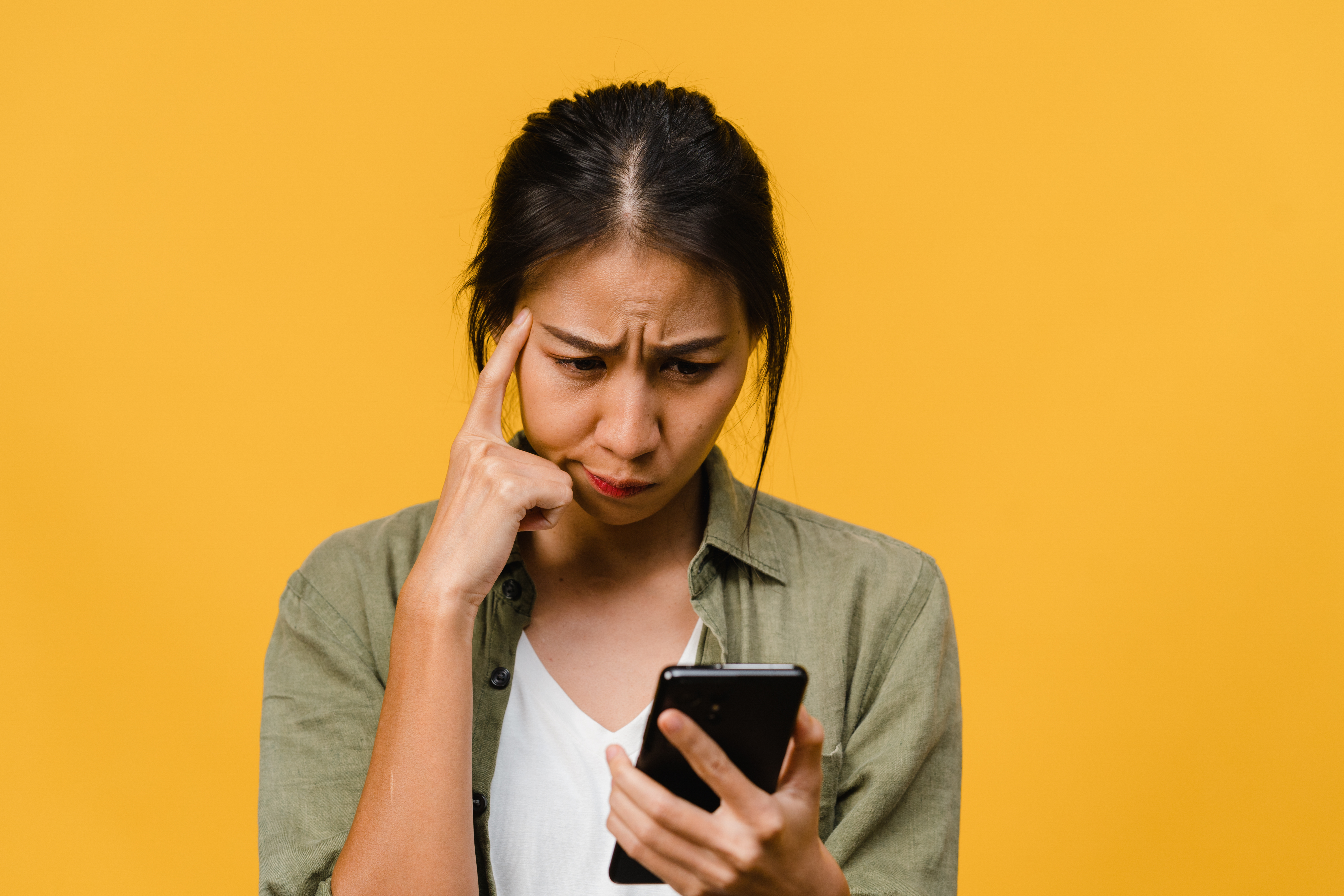 Uma jovem asiática sobre fundo amarelo olhando para celular com expressão pensativa sobre o que é cash out e quando encerrar uma aposta que fez