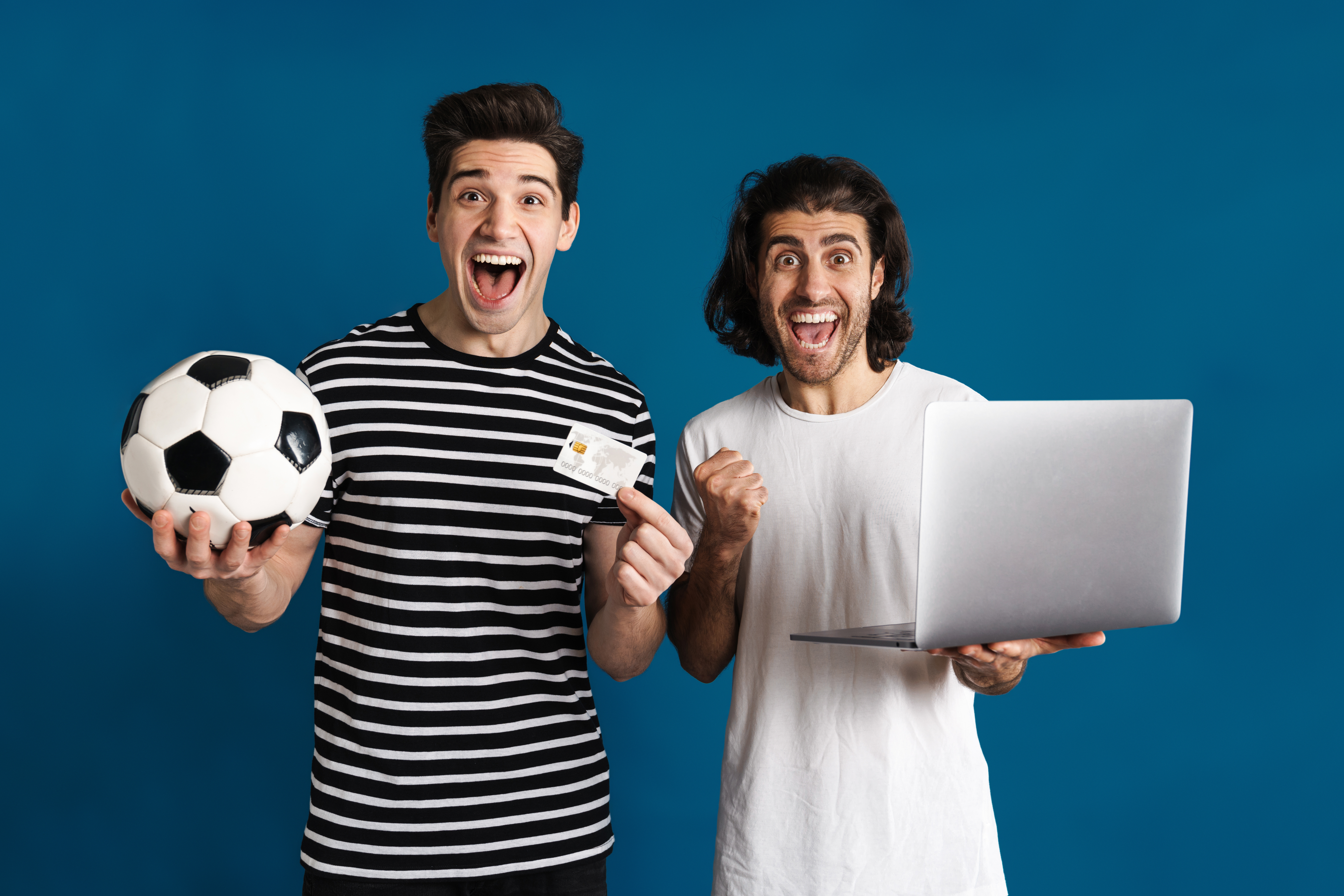 Dois homens animados usando laptop enquanto seguram bola de futebol e cartão de crédito, buscando saber mais sobre o bolão da Novibet